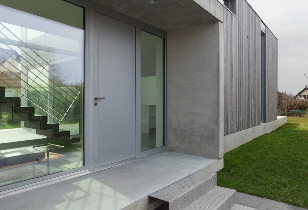 Szare aluminiowe drzwi zewnętrzne obok dużego przeszklenia na zewnątrz szarego domu